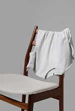 Weißes T-Shirt von KRONSTADT über Holzstuhl mit hellem Stoffsitz, vor grauem Hintergrund